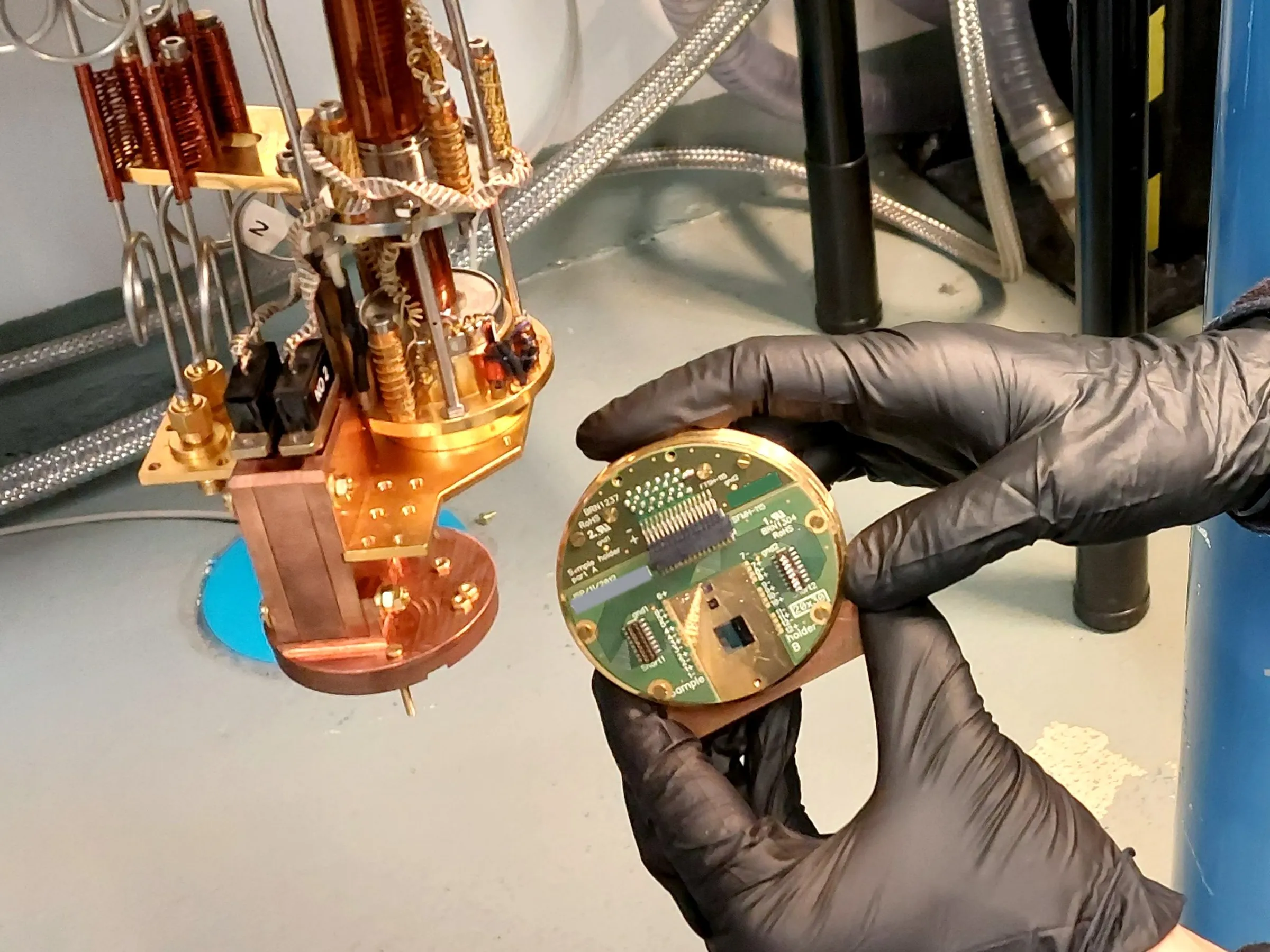 black-gloved-hands-hold-a-circular-circuit-board-near-a-contraption-of-narrow-pipes-and-brass-colored-plates.webp