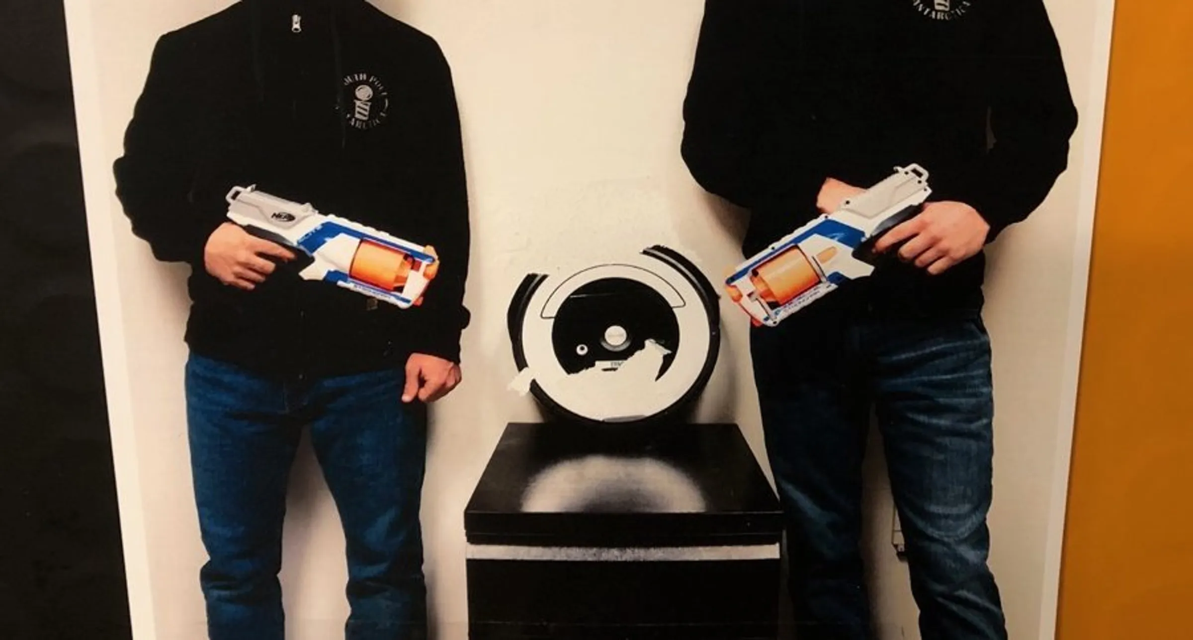 a-photo-of-two-men-from-the-shoulders-down-standing-and-pointing-nerf-guns-at-a-roomba-on-a-pedestal-between-them.webp