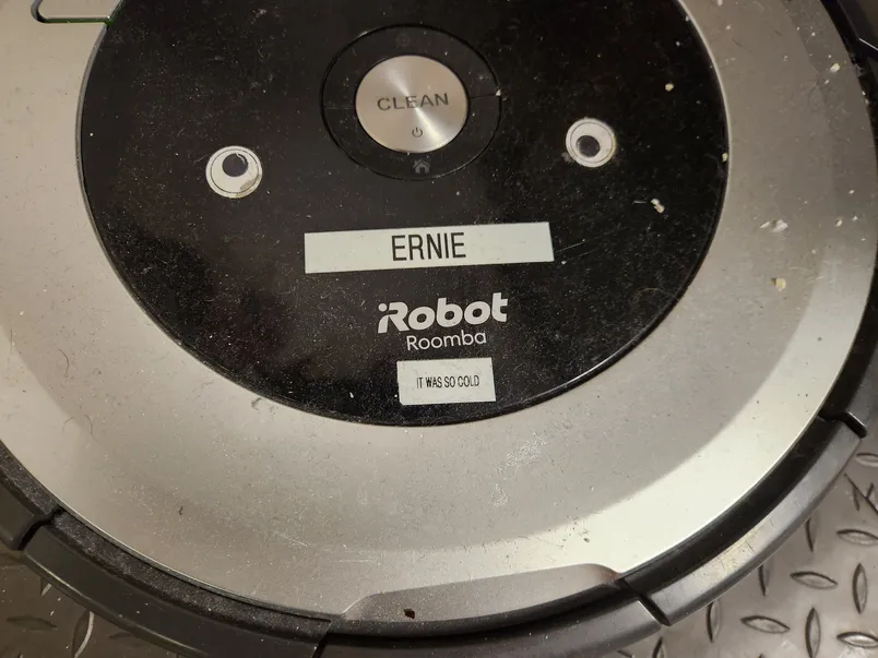 a-close-up-picture-of-the-top-of-a-roomba-showing-some-small-eye-stickers-a-sticker-with-the-name-ernie-and-a-sticker-that.webp