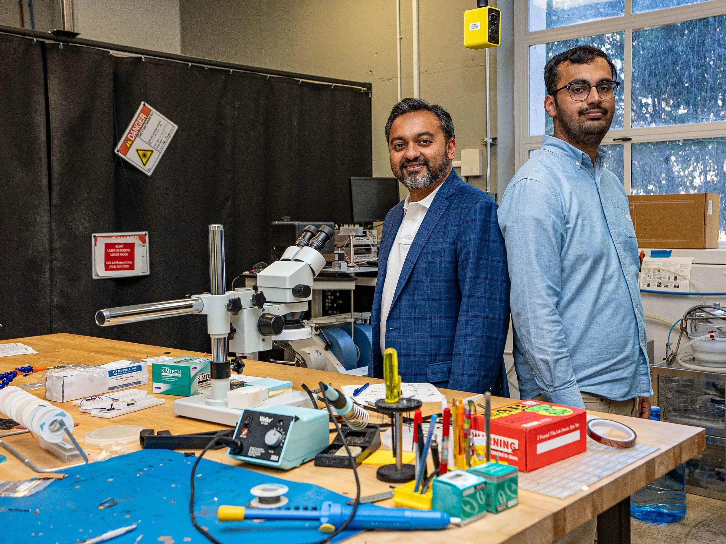 two-men-posing-for-a-portrait-with-electronics-on-a-table-in-front-of-them.webp